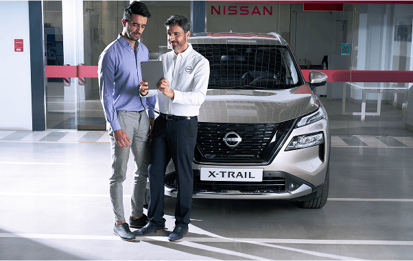 Zwei Männer stehen vor einem silbernen Nissan X-Trail, einer zeigt dem anderen etwas auf einem Tablet in einem Autohaus.