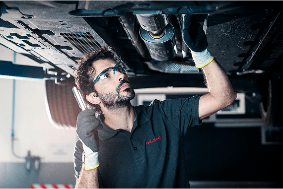 Ein Mechaniker von Nissan mit Schutzbrille und Handschuhen inspiziert die Fahrzeugunterseite eines Fahrzeugs mit einer Taschenlampe in einer Werkstatt.